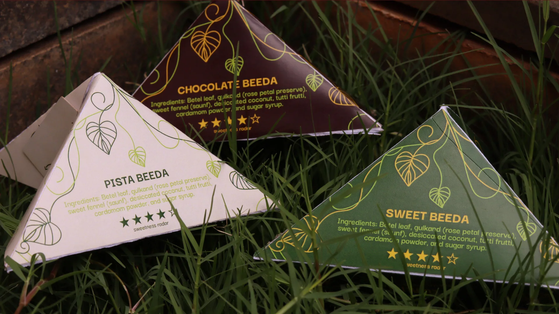 Three pyramid beeda packs on grass: Pista in cream, Chocolate in maroon and Sweet in green, each with gold leaf line-art and its ingredients