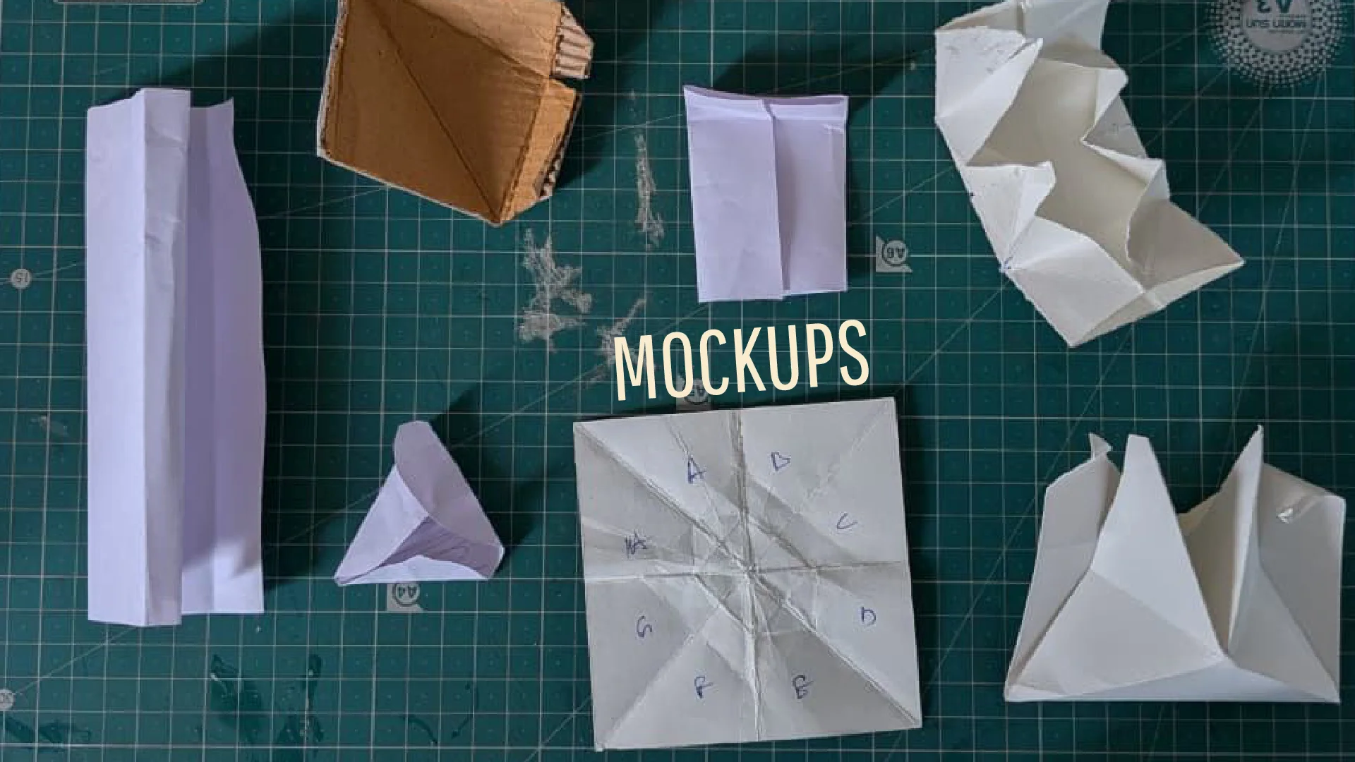Paper mock-ups on a cutting mat: pouches, a cone, a crumpled wrap and pyramids