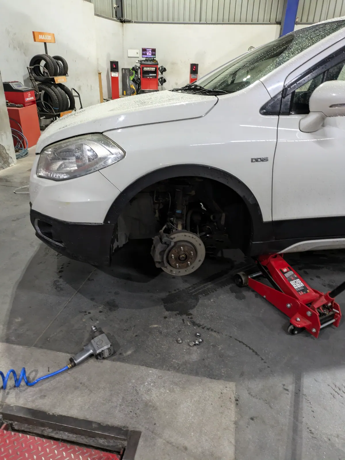 A white car in a tyre shop on a trolley jack, its front wheel off, an impact wrench on the floor